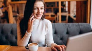 A woman with long dark hair is sitting in a cozy café, speaking on her phone and working on a laptop. She is wearing a white turtleneck sweater and smiling. In front of her is a cup of coffee on the wooden table. The background shows a wooden partition.