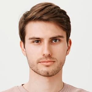 A young man with short, brown hair and a light beard is staring directly at the camera with a neutral expression. He is wearing a light-colored shirt, and the background is plain and light-colored.