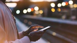 A person, partially visible from the side, is using a smartphone at a train station. The background shows a blurred view of the tracks and bright bokeh lights, conveying an evening or night setting. The person is wearing a light-colored jacket.