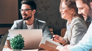 Three people are sitting at a table, engaged in a lively discussion. Two are using laptops, and the third is holding an open book. All are smiling and appear to be in a positive and collaborative work environment. A small potted plant sits on the table.