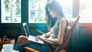 A woman sits in a cafe by large windows, intently working on her laptop. She is wearing a sleeveless top and jeans, with sunlight streaming through the window. There are two cups on a small table beside her.