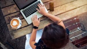 A person is seated outdoors at a wooden table, typing on a laptop. A smartphone lies next to the laptop, and a cup of coffee with latte art is placed on the left side of the laptop. The person wears a smartwatch on their left wrist.