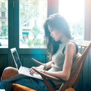 A woman with long dark hair sits on a wooden chair by large windows, intently working on a laptop placed on her lap. She wears a sleeveless top and jeans, and sunlight streams through the windows, creating a bright, airy atmosphere.