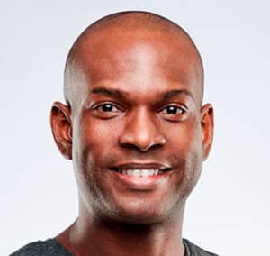 A smiling individual with a shaved head is pictured against a light background. They are wearing a dark-colored shirt and are looking directly at the camera.