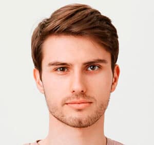 A young man with short brown hair and a trimmed beard is looking directly at the camera. He is wearing a light-colored shirt and has a neutral expression on his face. The background is plain and light.