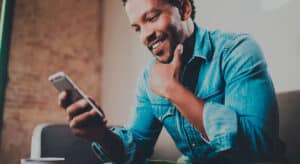 A person wearing a denim shirt sits on a couch, smiling while looking at a smartphone. One hand rests on their chin thoughtfully. The background is softly blurred, focusing attention on the individual.