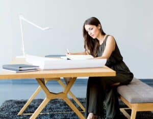 A woman sits at a modern wooden desk, writing on a large sheet of paper. She is focused and wears a dark green sleeveless dress. The desk holds books, a lamp, and more large sheets of paper. The setting is a bright, minimalistic office with grey floors and a textured rug.