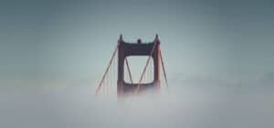 Image shows the top portion of a bridge, partially obscured by fog. The bridge features prominent vertical towers and suspension cables, with a hazy sky in the background. The scene exudes a mystical atmosphere due to the enveloping fog.