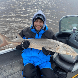 A person wearing a blue and black winter jacket and a gray beanie sits in a boat, smiling while holding a large fish with a pointed snout. The boat is on a body of water, which has small waves visible in the background.