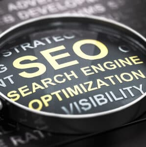 A close-up shot of a magnifying glass highlighting the words "SEO Search Engine Optimization" in yellow text on a black background, with other related, less focused terms like "Visibility" and "Strategy" visible around the main text.