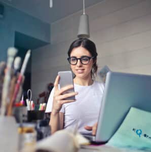 A person wearing glasses and a white shirt sits at a desk, holding a smartphone and smiling. In front of them is an open laptop. Art supplies, including paintbrushes, are scattered on the desk. A ceiling light hangs above.