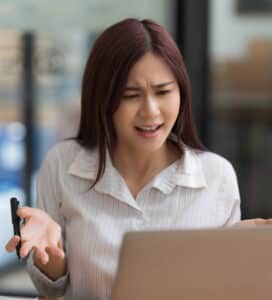 A woman with long, dark hair is sitting in front of a laptop, looking frustrated. She is wearing a striped shirt and holding a pen in her right hand while her left hand is raised in a questioning gesture. The background is blurred with indoor elements.