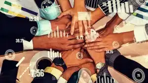 A group of diverse people place their hands together in a show of unity at home. Various data overlay elements, including graphs, numbers, and progress bars, are positioned around the hands, indicating metrics and analytics. A cup of coffee and a smartwatch are visible.