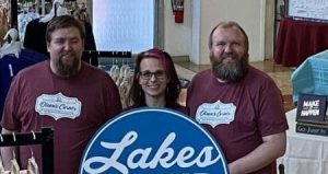 Three people stand inside a store, smiling at the camera. They are wearing matching maroon t-shirts. In front of them is a large circular sign that reads "Lakes Proud, Shop. Buy. Support." Clothing items are displayed around them, highlighting the services they offer both in-store and on their website.