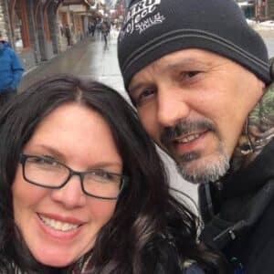 A smiling woman with long dark hair and glasses stands next to a man with a goatee and a beanie. They are outdoors on a rainy street with wet pavement, surrounded by buildings, capturing a cozy sense of home amidst the urban setting.