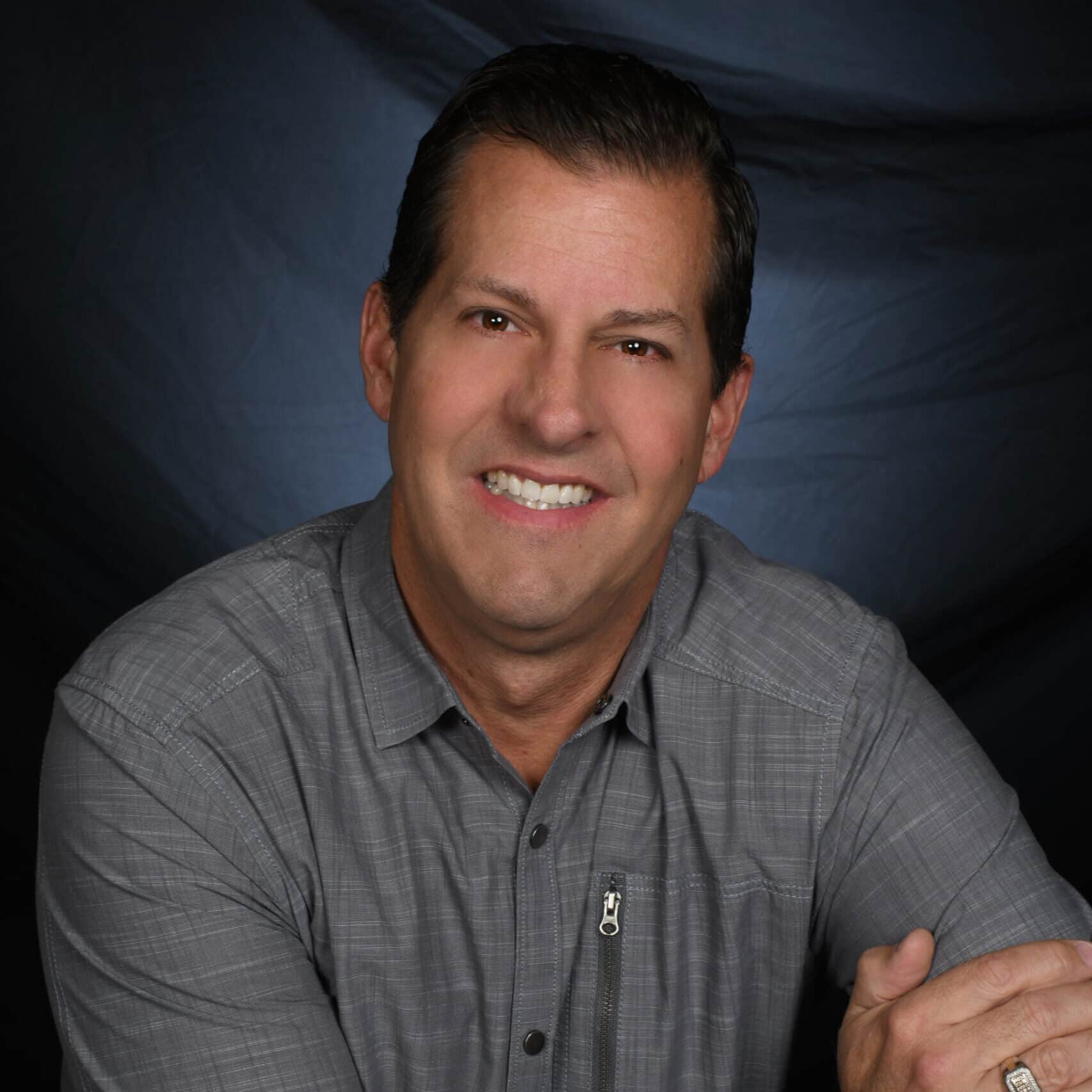 A person with short dark hair and smiling is posed against a dark backdrop. They are wearing a grey button-up shirt and have their arms resting in front of them, ready to share client testimonials.