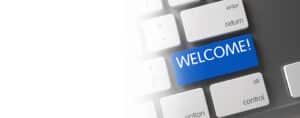 A keyboard with a blue key labeled "WELCOME!" in place of the usual "Enter" key. The surrounding keys are white with standard labels like "return" and "control." The left side of the image fades into a white background, giving it a clean and home-like aesthetic.