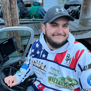 A smiling man sits at the helm of a boat docked by a wooden pier. He is wearing a gray baseball cap, a hoodie with various logos and an American flag design, and is gripping the boat's steering wheel.