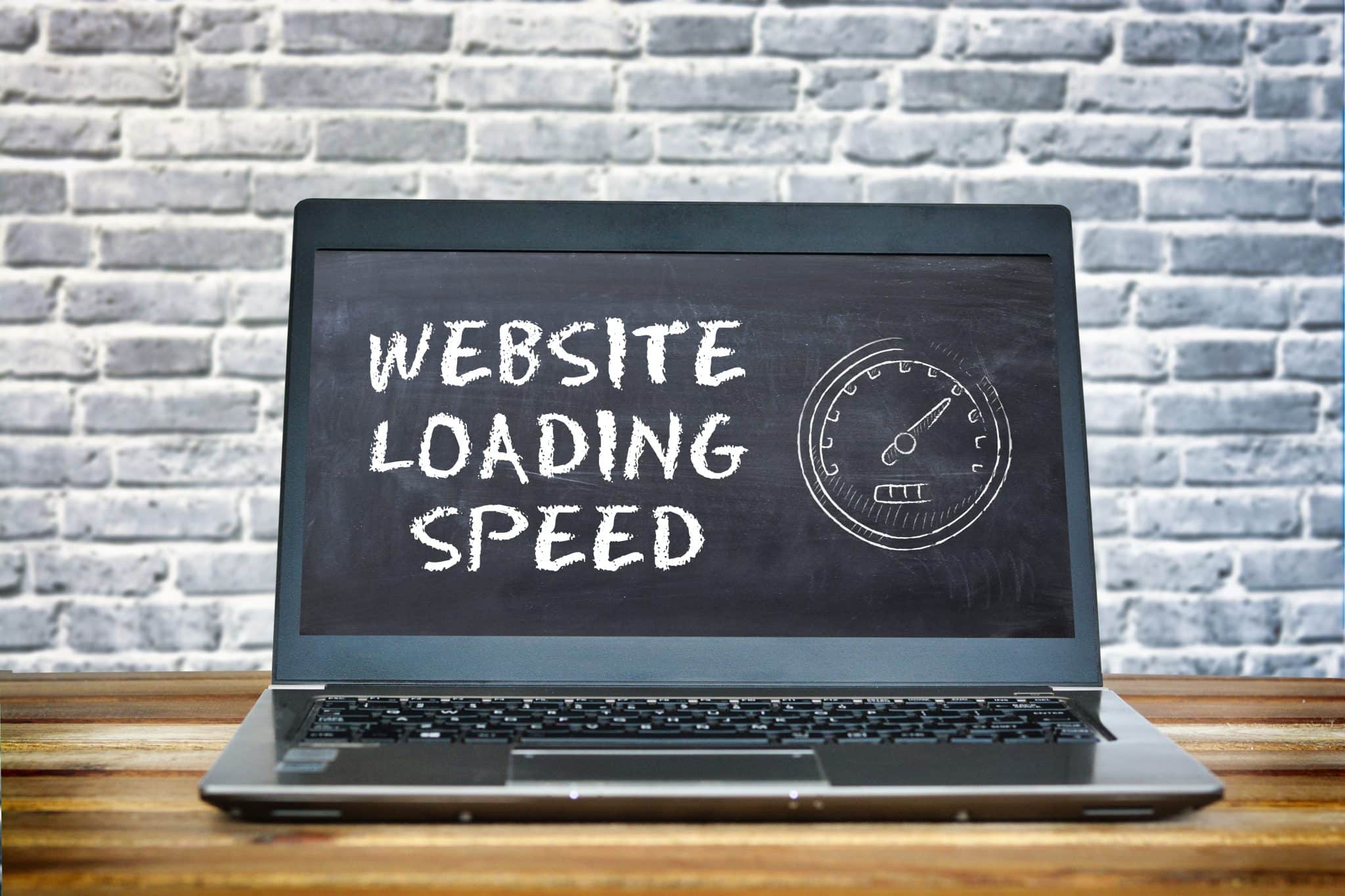 A laptop on a wooden table displays the text "Website Loading Speed," emphasizing its importance, on a screen resembling a chalkboard. A speedometer icon is also shown. The background features a white brick wall, highlighting the significance of website performance.