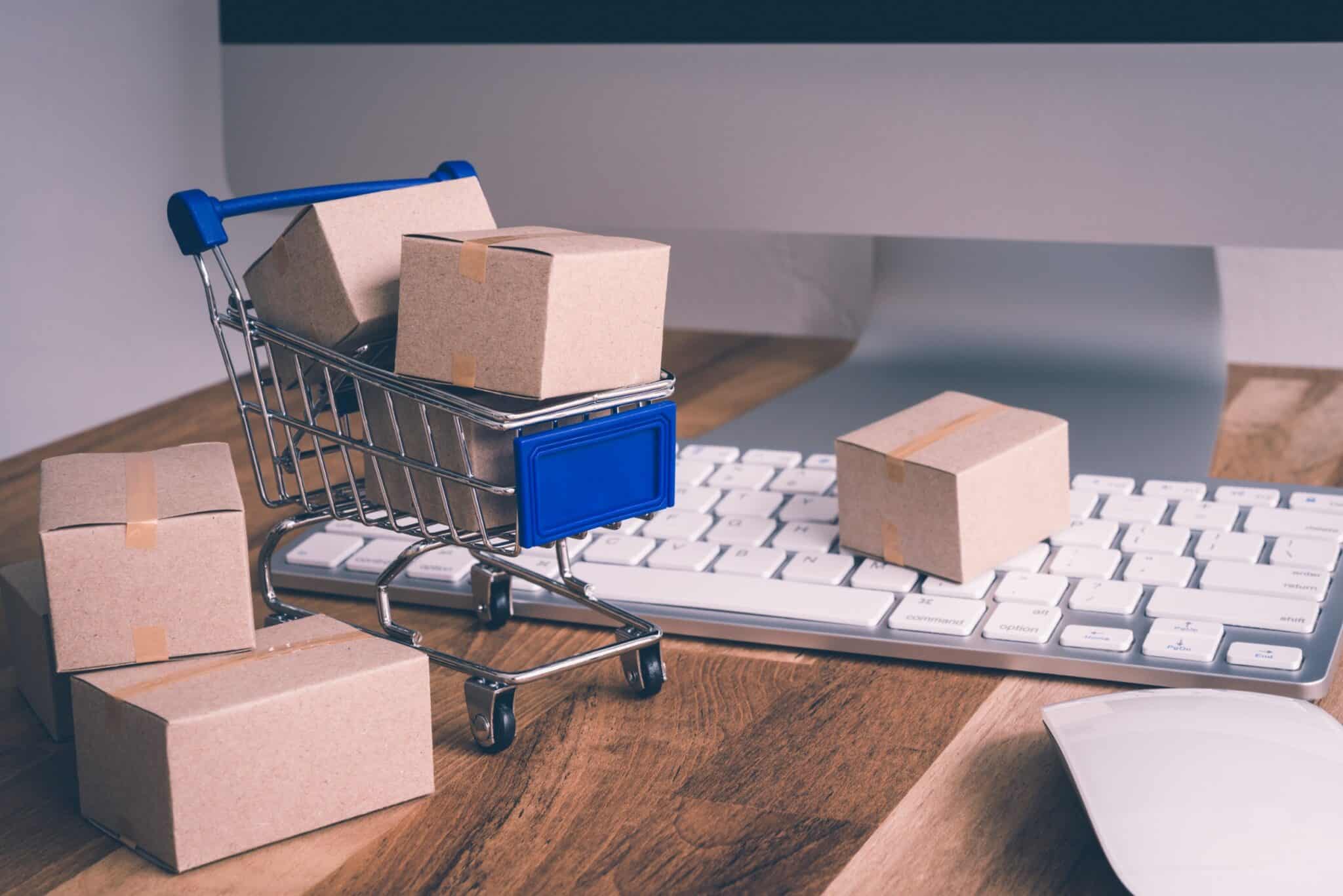 A miniature shopping cart filled with small cardboard boxes sits on a computer keyboard, with additional boxes around it. The scene, set on a wooden desk with a computer mouse and part of a monitor visible, vividly suggests the user-friendly nature of e-commerce websites.