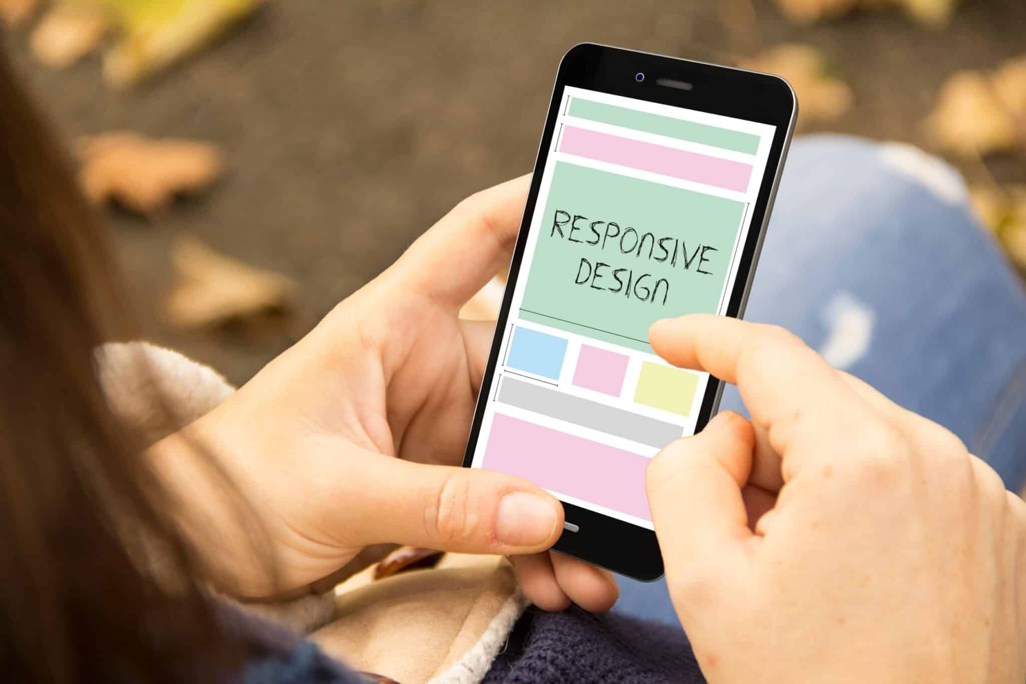 A person is holding a smartphone, their index finger touching the screen. The display features a webpage layout with "Responsive Design" prominently centered. In the background, leaves are scattered on the ground, giving an organic feel to this seamless auto draft experience.