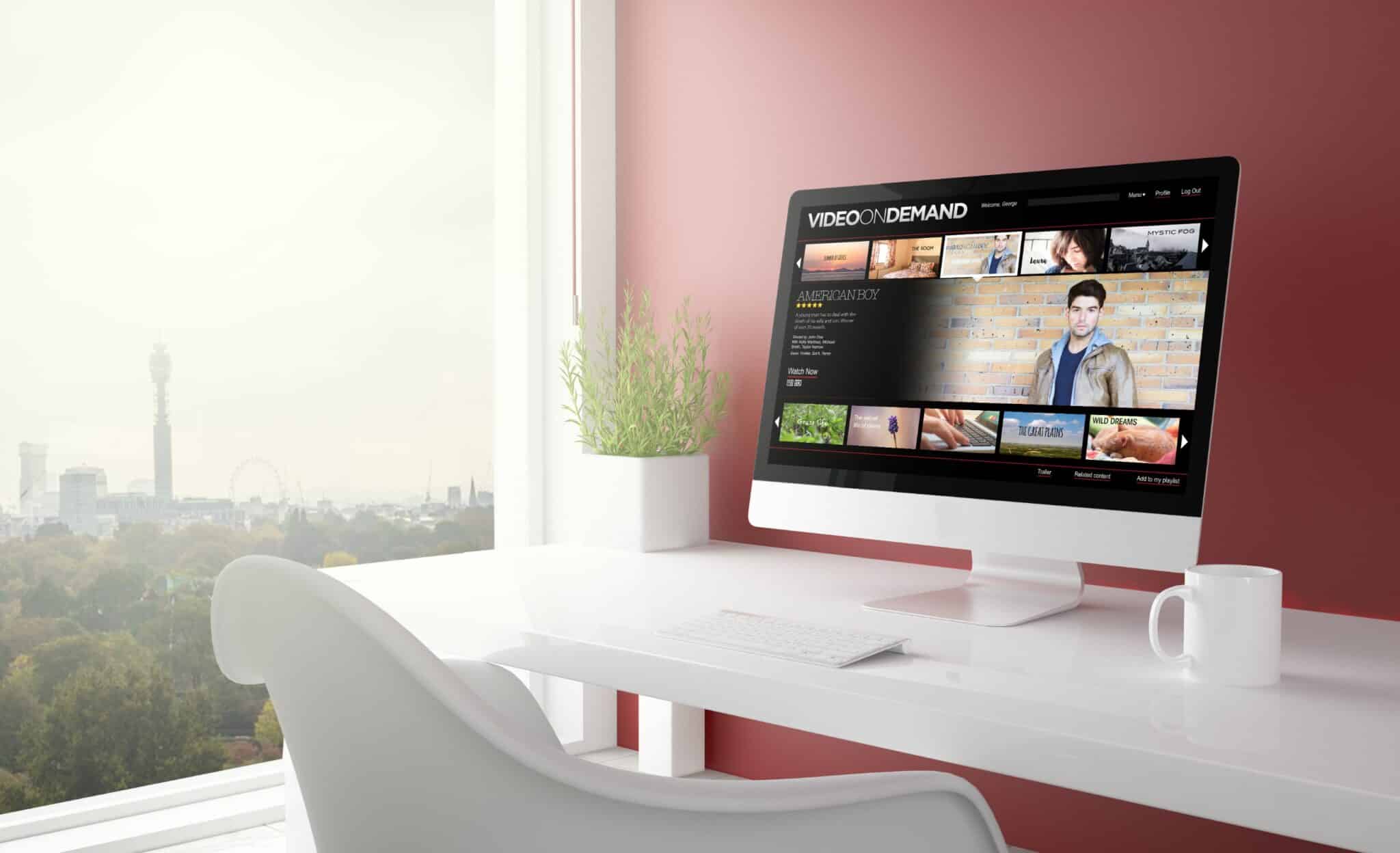 A modern workspace with a white desk and chair placed by a large window. A computer on the desk displays an auto draft of a video-on-demand service. A potted plant also adorns the desk. The window offers a view of a cityscape with the London skyline, featuring the London Eye and BT Tower.