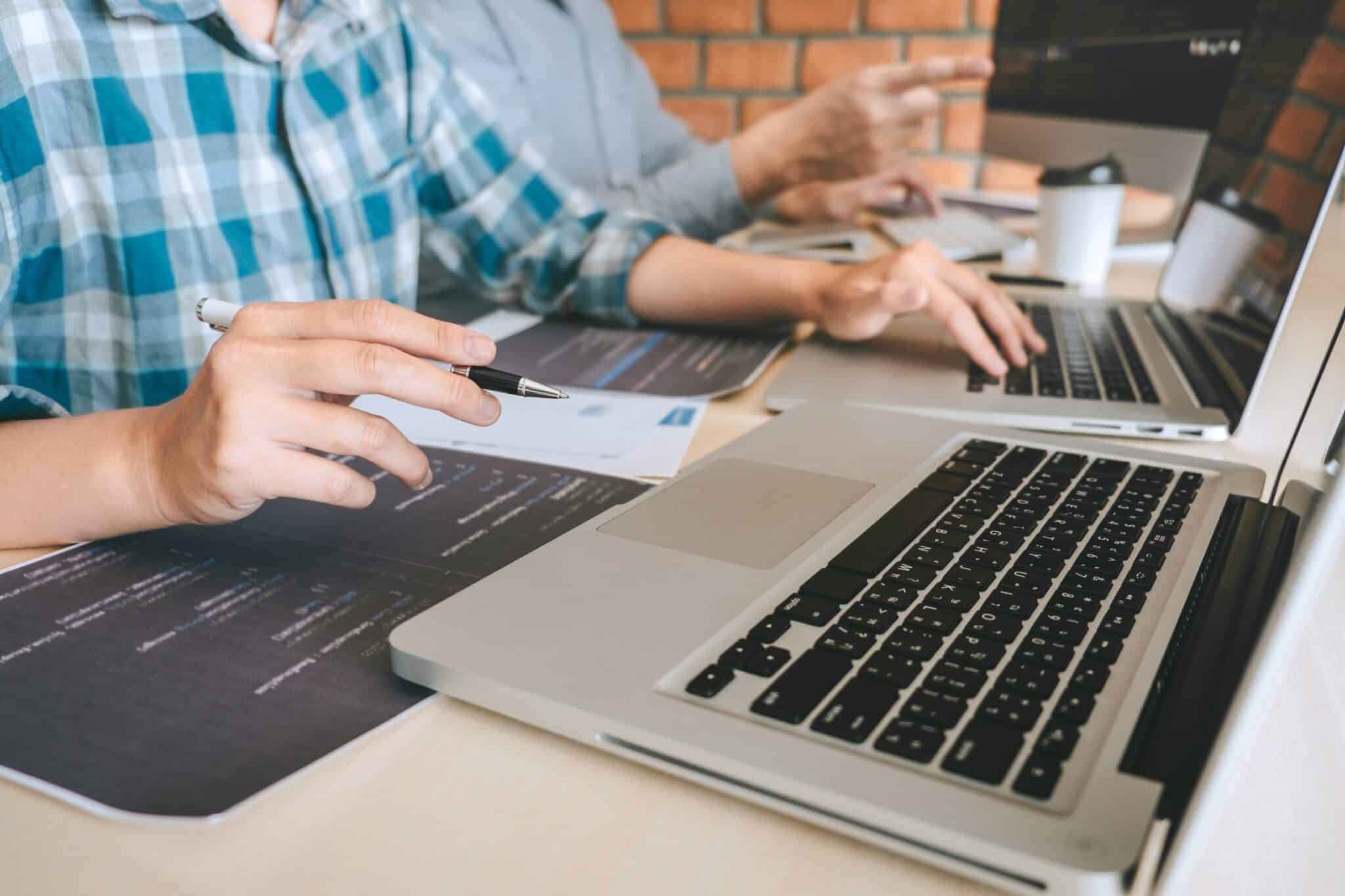 Two people sit at a table with laptops open, focused on screens. One person points at a monitor while holding a pen, emphasizing the crucial discussion on business strategies. A coffee cup sits in the background, hinting at a long session of collaborative work on website accessibility.