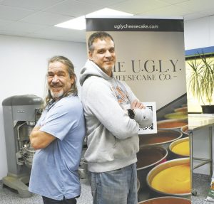 Two men standing back-to-back, both smiling. One is wearing a light blue shirt, and the other is in a gray hoodie. Behind them is a banner that reads "The U.G.L.Y. Cheesecake Co." with images of cheesecakes.