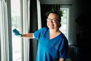 A smiling person in blue scrubs and glasses is holding a cloth by a large window. They appear to be cleaning. The room is well-lit with natural light coming through the window.