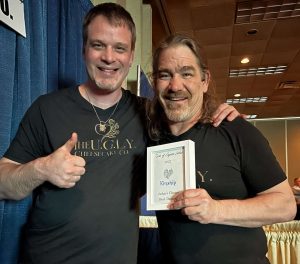 Two smiling men stand together indoors. The man on the left gives a thumbs up and wears a shirt with "THE UGLY JERKY" on it. The man on the right holds a certificate labeled "Award of Excellence" and wears a matching shirt.