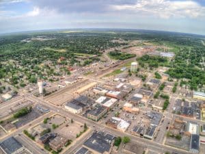 Aerial view of a small town with grid-like streets and a mix of buildings, roads, and greenery. Two water towers are visible, and a railway line runs through the area. The horizon shows vast expanses of green landscape under a partly cloudy sky.