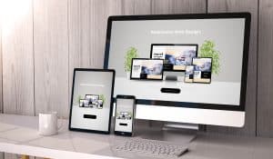 A computer monitor, tablet, and smartphone displaying a responsive web design on a wooden desk with a white coffee mug and notebook beside them. The backdrop features a wooden wall and green plants.