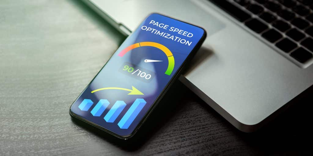 A smartphone showcasing website speed optimization with a gauge reading 90/100 sits next to a laptop. A bar graph enhances the display below the gauge, highlighting SEO improvements. The phone rests on a wooden surface.