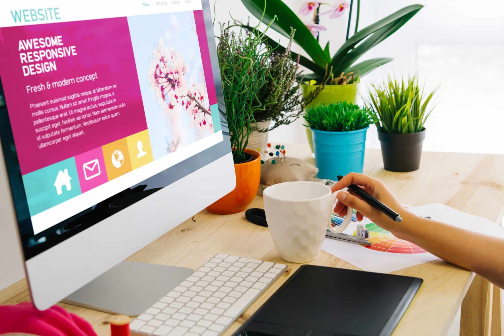 A person at a desk with a computer showcasing a colorful website design is focused on enhancing user experience. The desk holds potted plants, a white mug, a color palette, and a keyboard and pen. The screen displays text about responsive design with vibrant icons and sleek typography.