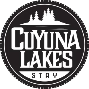 A circular logo with the text "Cuyuna Lakes Stay" in bold letters. Above the text, there are silhouettes of trees and a lake, all in white against a black background.