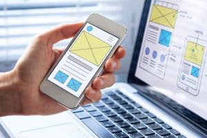 A person holds a smartphone displaying a wireframe design. In the background, a laptop screen shows a similar design schematic for a mobile app by the best website designer. Brightly lit workspace with a focus on app development and user interface design.