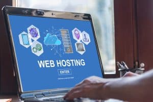 A laptop with a blue screen displays the text "WEB HOSTING ENTER 2025" and various tech icons, including a cloud, network symbols, and server graphics. In a sunlit room, hands poised on the keyboard emphasize the business-ready environment.