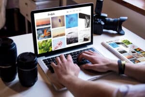 A person types on a laptop displaying a photography portfolio website, surrounded by a camera, two camera lenses, and a printed portfolio booklet—perfect inspiration to redesign your website by the window.