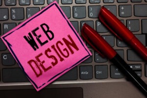 A pink sticky note with "WEB DESIGN" in bold black and red letters sits on a laptop keyboard next to two red-and-black pens, highlighting the importance of professional web design for small businesses.