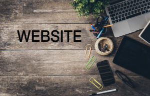 A wooden desk with a laptop, tablet, smartphone, stationery, paper clips, a small plant, and the word "WEBSITE" written in bold black letters on the left side.