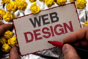 A person holds a card reading "WEB DESIGN" in bold letters, surrounded by crumpled yellow paper balls on a metallic surface, while marking the card with a red pen.
