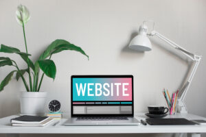 A laptop displaying the word "WEBSITE" sits on a white desk with a potted plant, desk lamp, cup, saucer, clock, notebook, and colored pencils—perfect for those ready to elevate your presence with a professional business web setup.