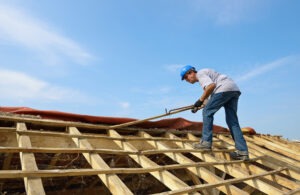 A construction worker in a blue hard hat and gloves stands on a wooden roof frame, showcasing the strength of local partner teamwork under a clear blue sky.