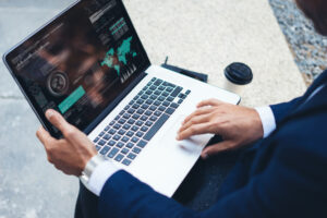 A person in a dark suit is using a laptop displaying data and graphs, likely exploring web design or marketing services to boost your business. A disposable coffee cup sits nearby on a stone surface in this outdoor scene.