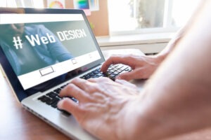 A person types on a laptop showing a screen with the words "#Web DESIGN" and icons of devices, suggesting work on web design. The scene is bright with natural light coming through a window.