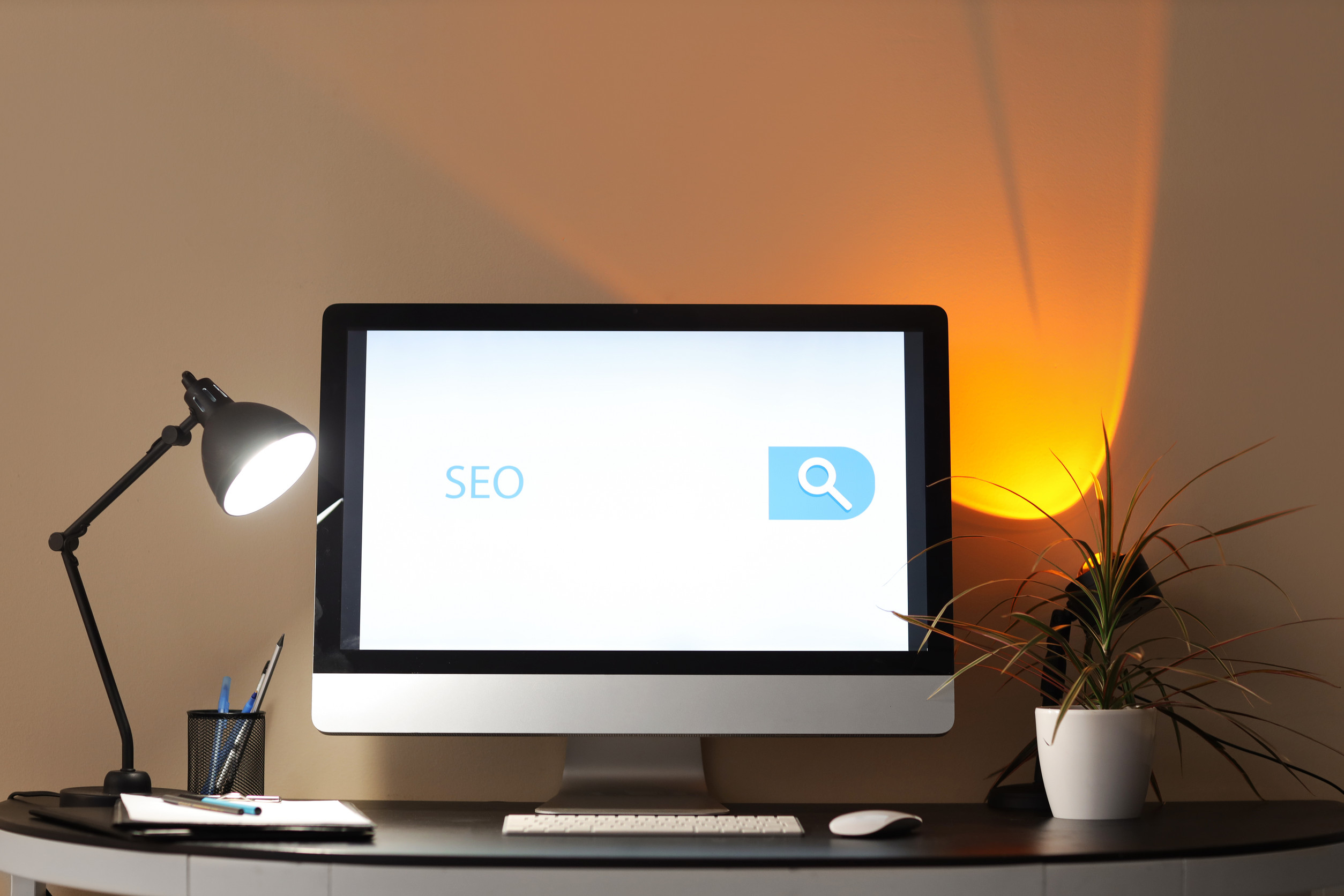 A desktop computer on a black desk displays the word "SEO" next to a search icon. A black desk lamp, a potted plant, pens in a holder, and a keyboard are also on the desk. A warm light glows on the wall behind.