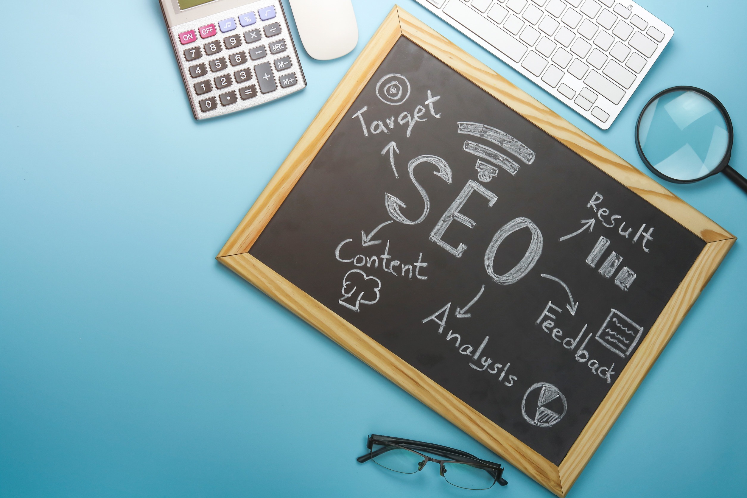 A chalkboard with "SEO" written at the center, surrounded by related terms like target, content, analysis, result, and feedback; nearby are a keyboard, calculator, magnifying glass, and glasses on a blue background.