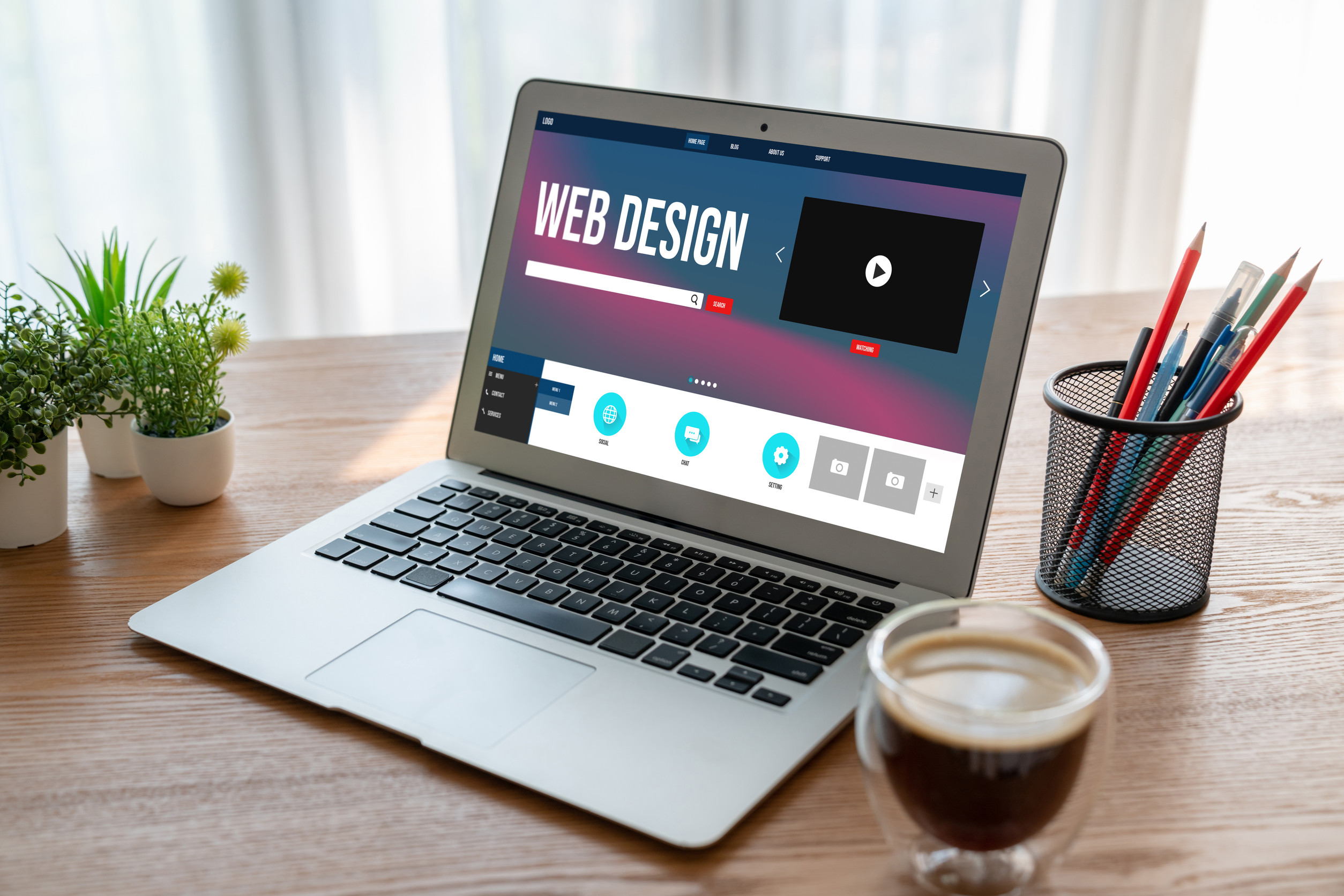 A laptop displaying a web design application sits on a wooden desk with a cup of coffee, a small plant, a container holding pens and pencils, and a window with sheer curtains in the background.