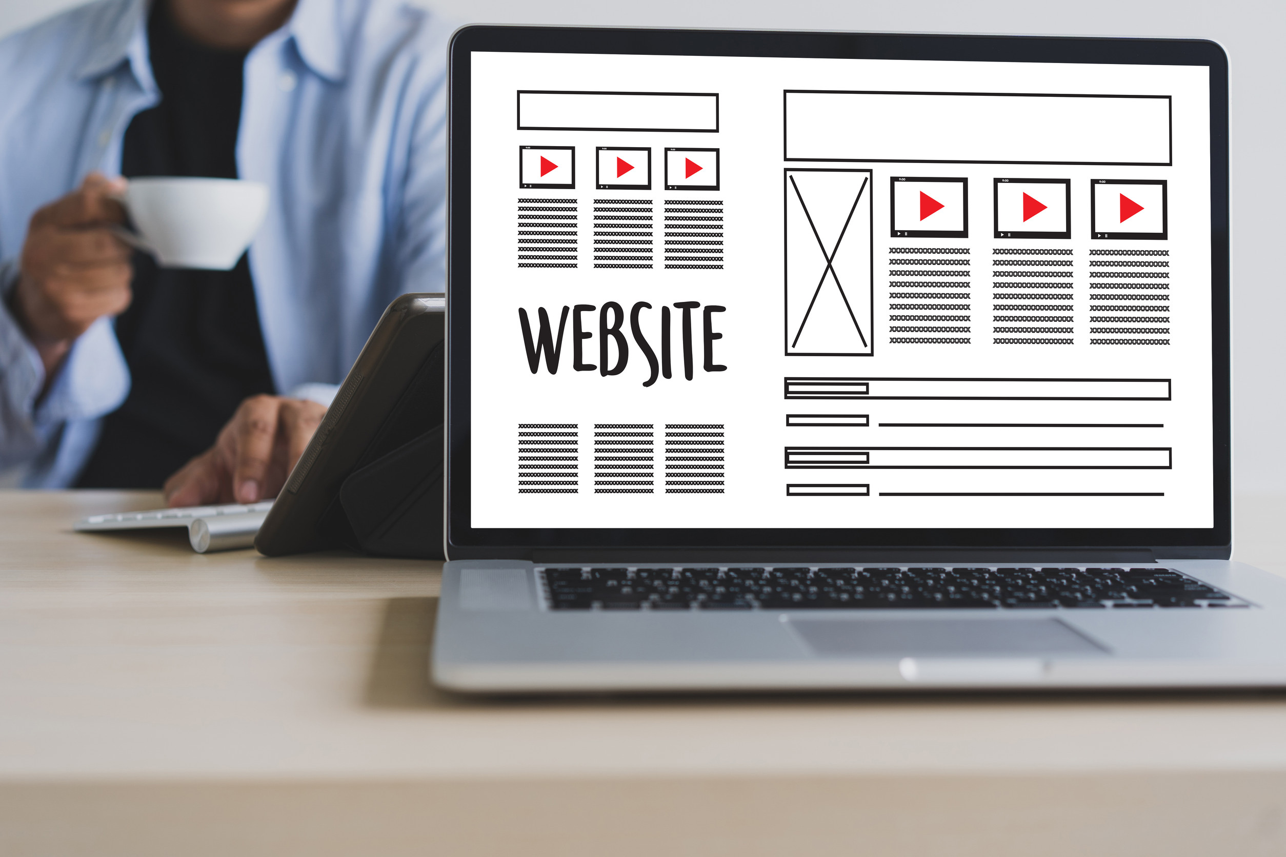 A laptop displays a website project wireframe with sections for videos, text, and images. In the background, a person sits at a desk holding a cup of coffee.