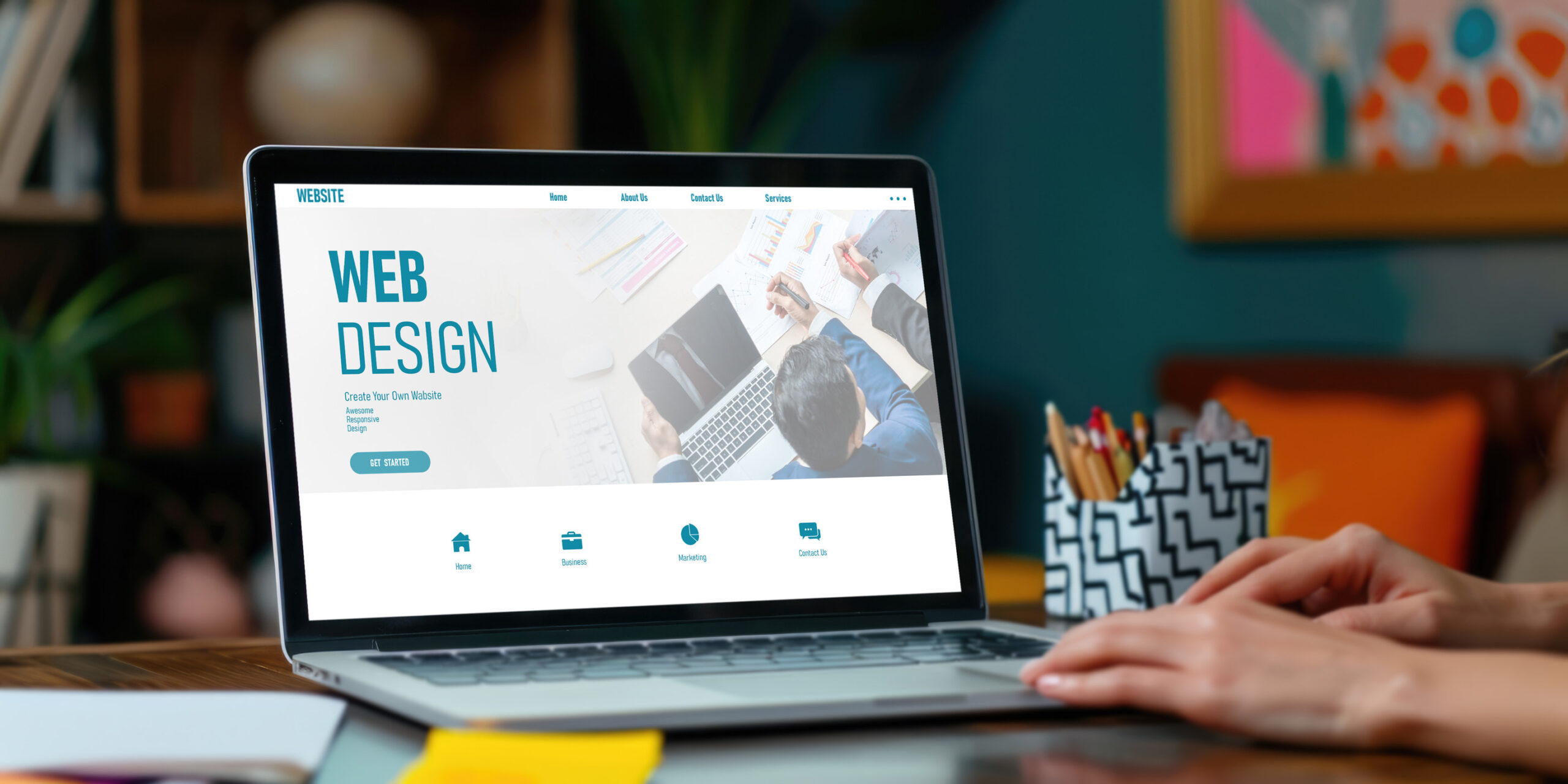 A person uses a laptop displaying a web design page with the heading "WEB DESIGN" and icons for various services. The workspace includes a pencil holder, sticky notes, and blurred background decor.